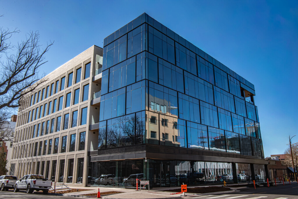 Modern office building with glass facade reflecting another structure, clear sky, and surrounding street elements